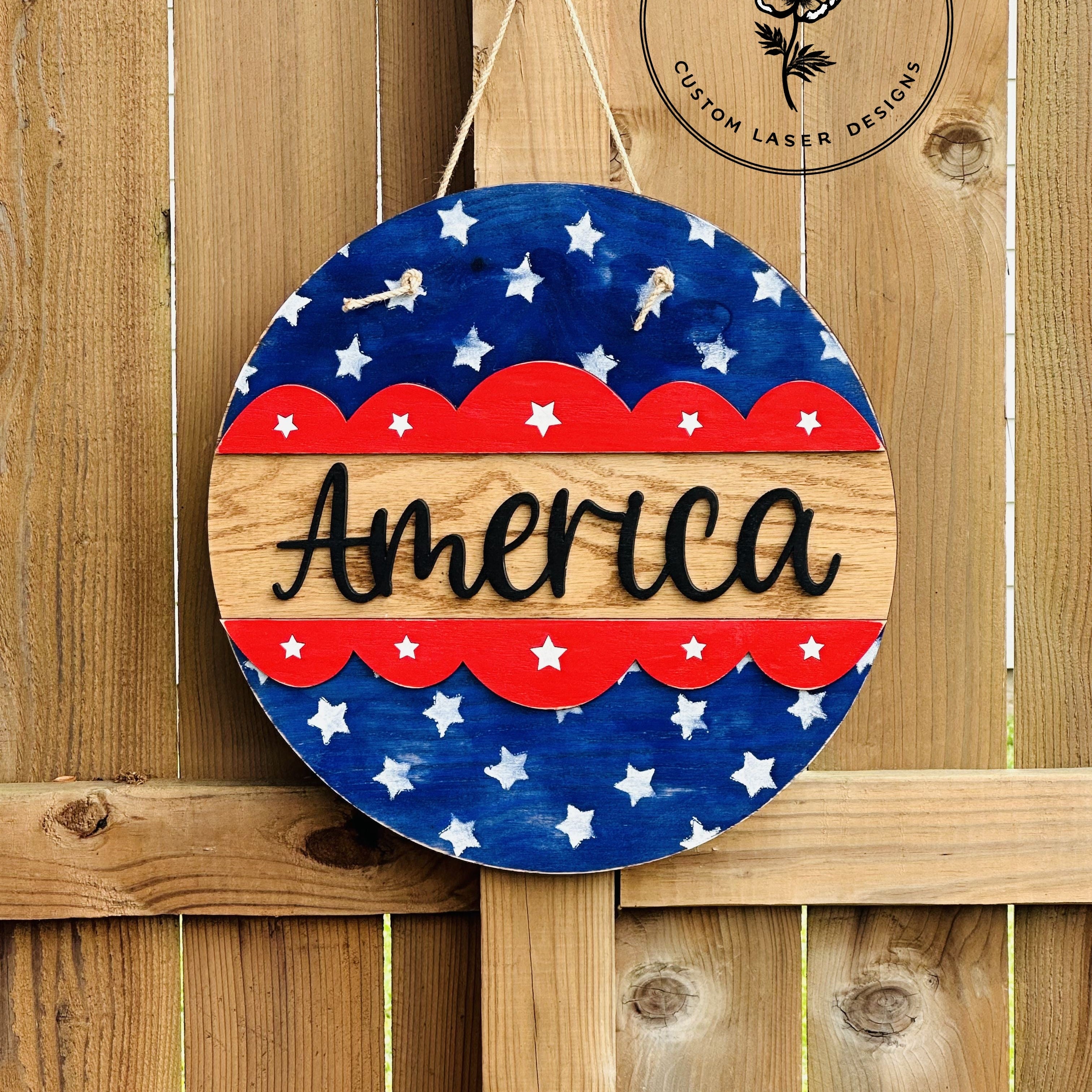 Round wooden sign with American flag design and 'America' text, hanging on a wooden fence with 'Wooden Wildflower' branding.