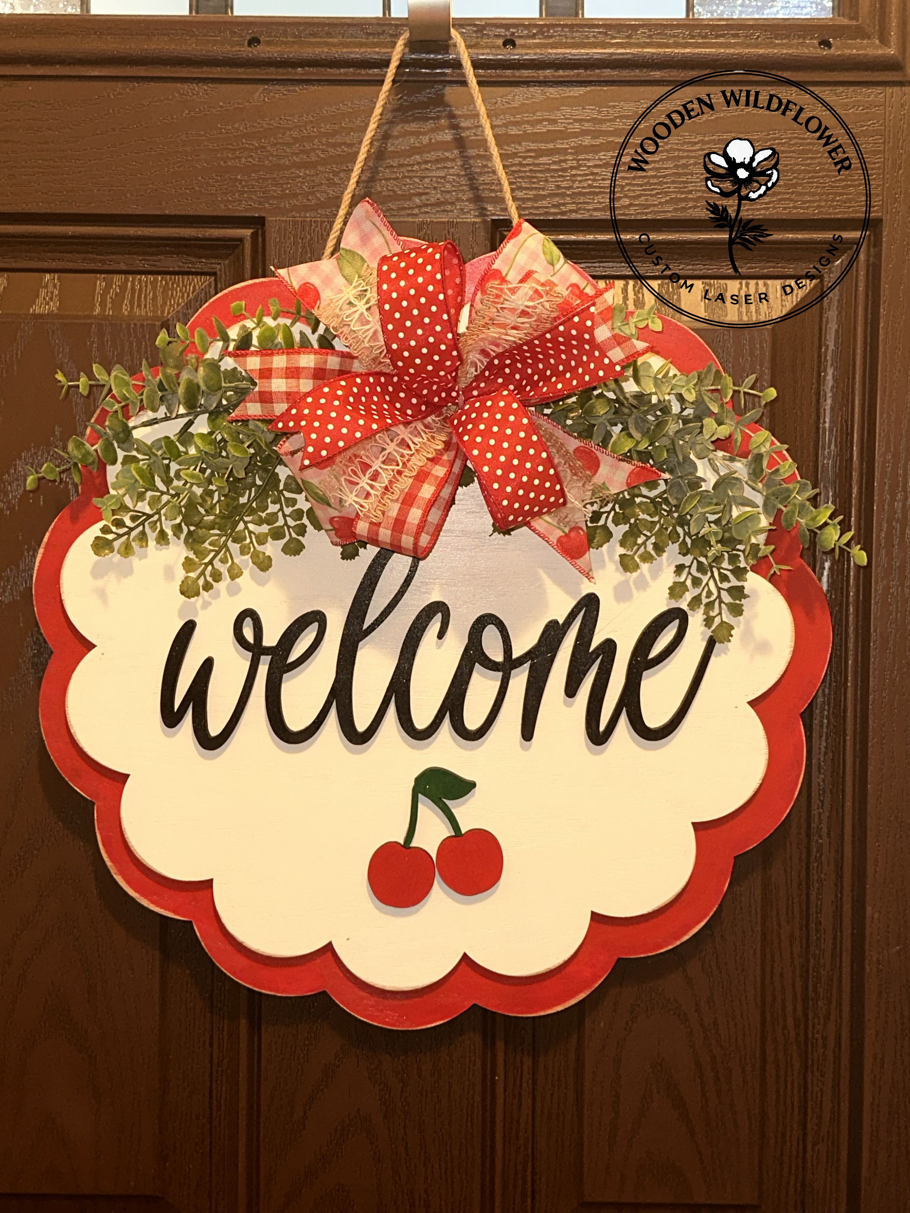 Decorative door hanger with a red bow, greenery, and 'welcome' text on a wooden background.