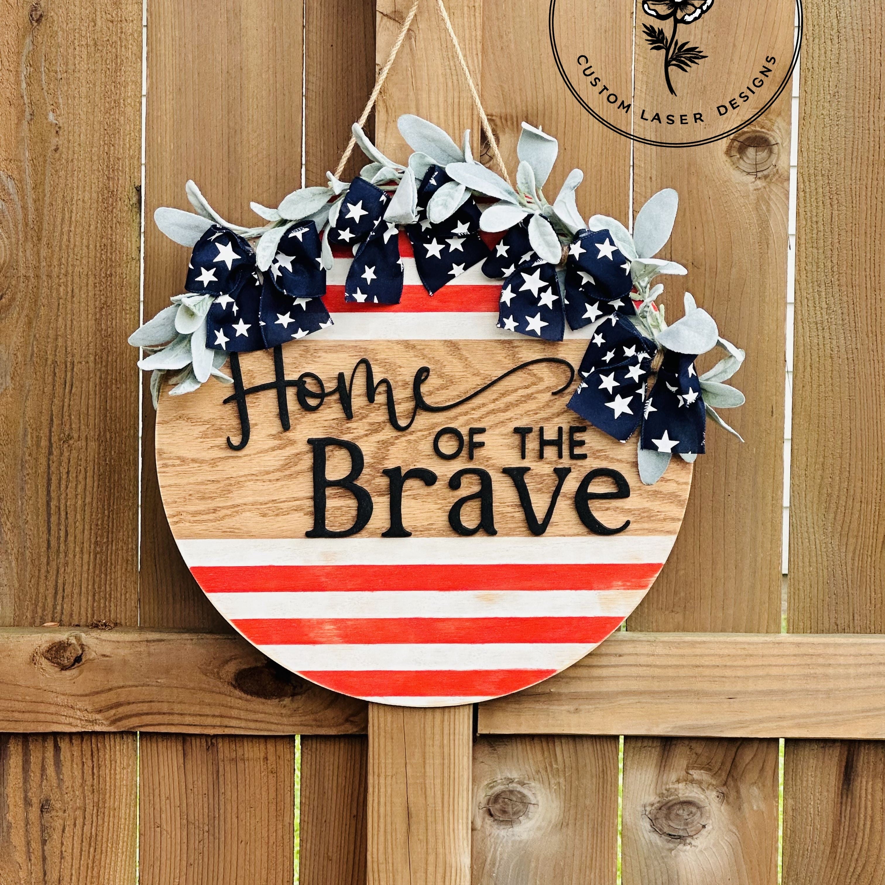 Decorative sign with American flag design and 'Home of the Brave' text, hanging on a wooden fence.